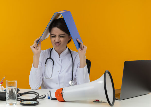 闭着坐在桌上的女听诊器和戴着黄色隔离眼镜的年轻医生笔记本电脑压力文件夹