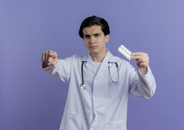 看皱眉的年轻男医生穿着医用长袍 听诊器显示一包药片 看着并指着紫色墙上的复制空间孤立皱眉显示年轻