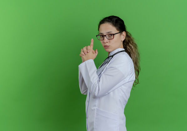 眼镜自信的年轻女医生 穿着医用长袍 戴着听诊器和眼镜 站在侧视图中 做着的姿势 隔离在绿色的墙上 留着复印空间医生站听诊器