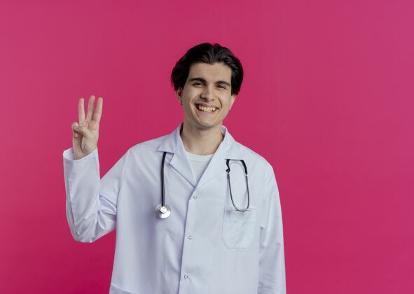 听诊器微笑的年轻男医生穿着医用长袍和听诊器 显示三只手被隔离在粉红色的墙上 并留有复印空间表演手男性