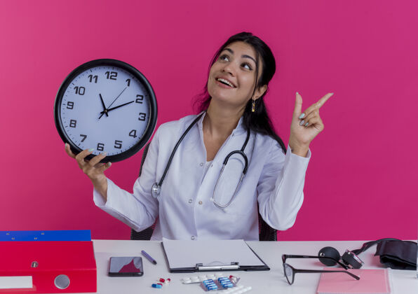 医疗令人印象深刻的年轻女医生穿着医用长袍和听诊器坐在办公桌旁 手持医疗工具 把头转向一边 抬起手指 孤立地站在粉红色的墙上长袍年轻人头