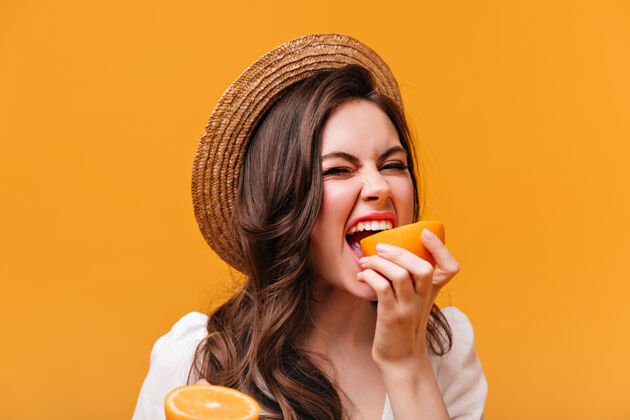 化妆戴着帽子的卷发女孩在孤立的背景下咬着多汁的橘子微笑船夫肖像