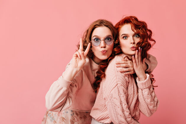 情绪欣喜若狂的欧洲女孩展示和平标志两位梦幻女郎在粉色背景下拥抱的摄影棚镜头时尚年轻人女性