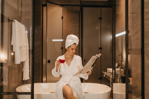 休息头上裹着毛巾 穿着浴衣的女人在看最新消息拿着一杯茶的女人在浴室里摆姿势身体浴室脸