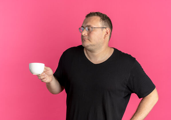 看戴眼镜的超重男子身穿黑色t恤 拿着咖啡杯 面带严肃的粉红色面容向一边看粉色拿着黑色