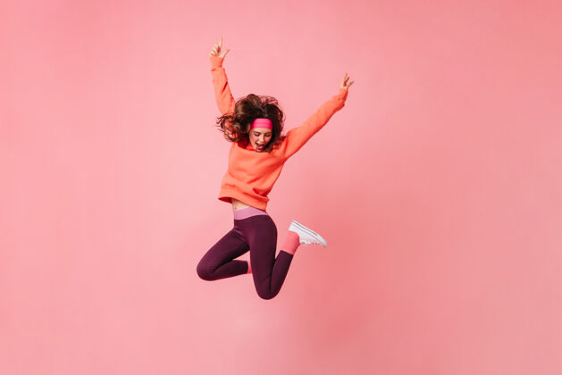 健美操可爱的黑发女子在粉红色的运动头带和运动服上跳孤立的粉红色墙壁头发女士运动衫
