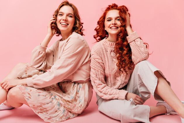 年轻人两个女孩微笑着坐在地板上在粉色背景上拍摄的朋友合影女孩欢呼成人