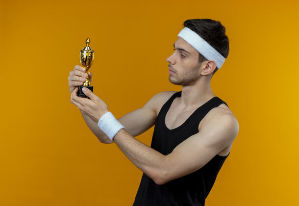 站着戴着头巾的年轻人拿着他的奖杯 带着自信严肃的表情站在橙色的墙上看着奖杯运动奖杯抱着