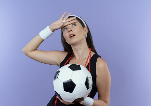 疲倦戴着头巾 脖子上系着跳绳 手里拿着足球 站在蓝色的墙壁上看着疲惫的年轻健身女士抱着脖子年轻