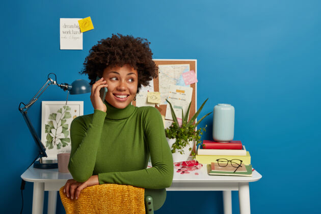 灯非裔美国妇女通过智能手机交谈 在家庭办公室办公桌上工作 有愉快的面部表情女人人手机