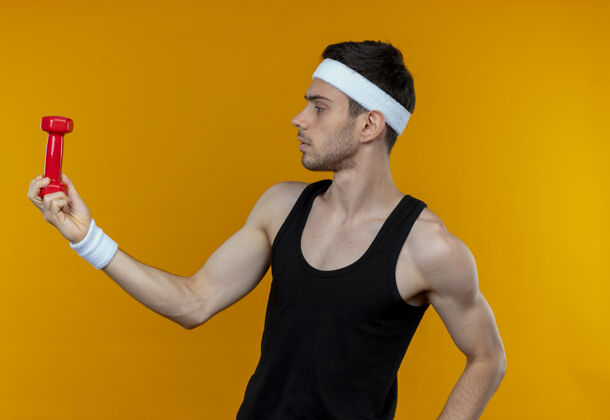 站戴着头巾 手持哑铃的年轻运动型男子站在橙色的墙上严肃地看着哑铃看哑铃年轻