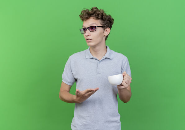 严肃戴着黑眼镜的年轻人 穿着灰色马球衫 手里拿着咖啡杯 一边看一边 脸上带着严肃自信的表情表情站着自信