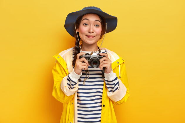深色一个长着长辫子 戴着帽子 穿着黄色雨衣 手持复古相机 微笑着的漂亮亚洲女人 在她令人敬畏的旅行中拍照 被隔离在黄色的墙上摄影条纹美女