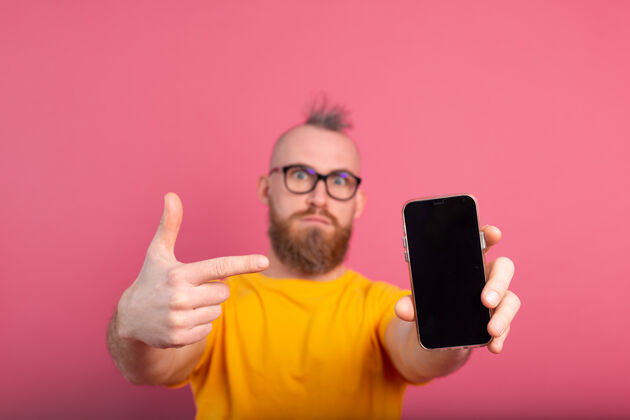 情感嘿 新的东西严重愤怒的欧洲胡子家伙指着他的手机与黑色的空白屏幕上的粉红色现代应用站着