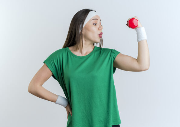 运动令人印象深刻的年轻运动女性戴着头带和手环举着哑铃手放在腰上看着她的肌肉保持头带抱着