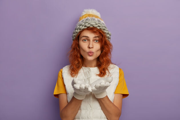 女性红发女人对着镜头吹 手掌向前 戴着针织帽子和白色背心 享受冬天的时光肖像亲吻横