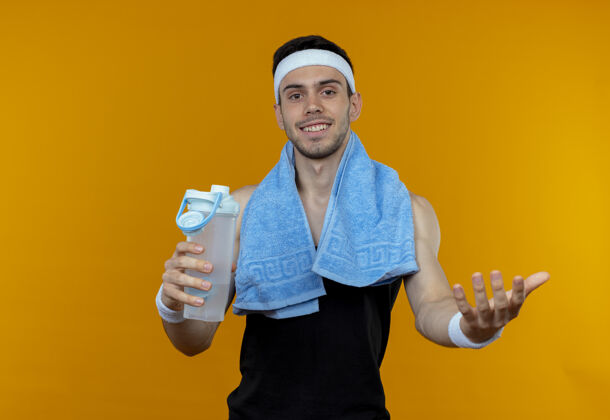 抱戴着头巾 脖子上围着毛巾 手里拿着一瓶水的运动型年轻人看着镜头 用橙色的手做着欢迎的手势水制造瓶子
