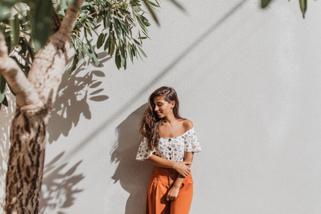 衬衫一位穿着橘色裤子和衬衫 光着肩膀的年轻女士倚在树下的白墙上 甜美地笑着深色头发快乐成人