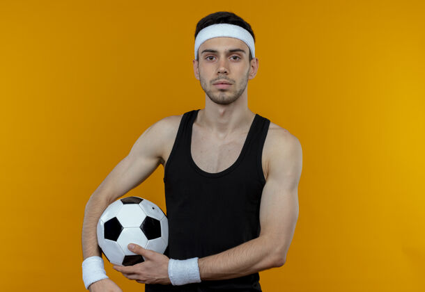 站着戴着头巾的年轻运动型男子拿着足球站在橙色的墙上 表情自信严肃男人头带球