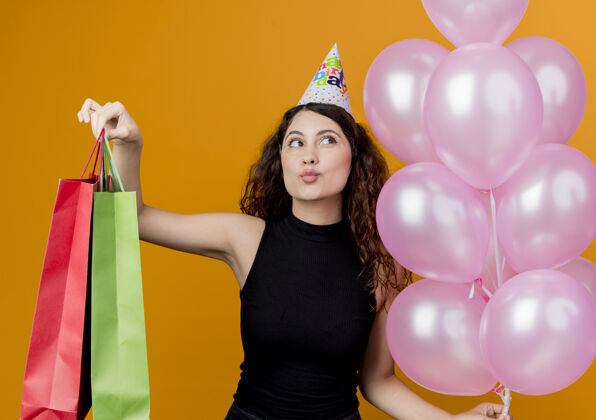 抱着一个年轻漂亮的女人 卷发 戴着节日帽 手里拿着空气和纸袋 站在橙色的墙上庆祝生日帽子包卷发