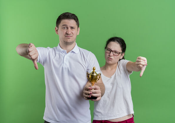 拇指一对年轻的运动情侣站在一起举着奖杯竖起大拇指站在绿色的墙上情侣男人女人