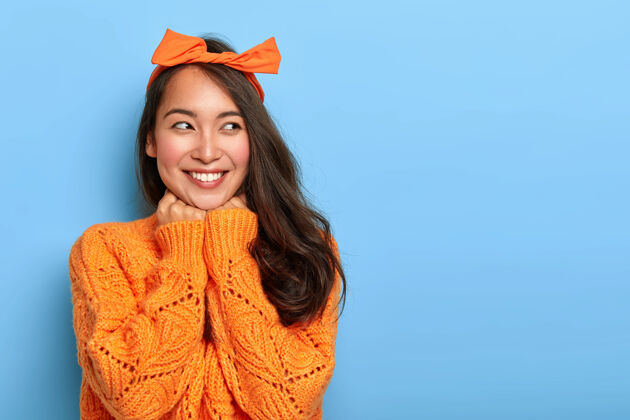 脸摄影棚拍摄的快乐乐观的女人穿着亮橙色针织套头衫 头戴蝴蝶结 下颚下牵手人类室内表情