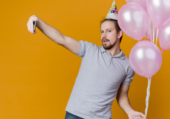 男人戴着节日帽的年轻人举着一堆气球在橙色的墙上用智能手机自拍庆祝生日束自拍使用
