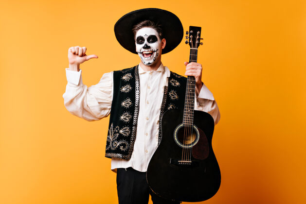 室内穿着墨西哥传统服装的兴奋的家伙拿着吉他快乐死歌手在万圣节派对上玩得开心情感恐怖万圣节