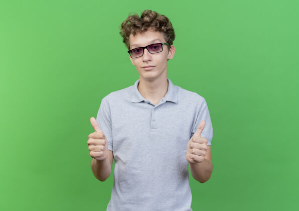 男人一个戴着黑眼镜 穿着灰色马球衫的年轻人站在绿色的墙边 愉快而积极地笑着竖起大拇指幸福站年轻人