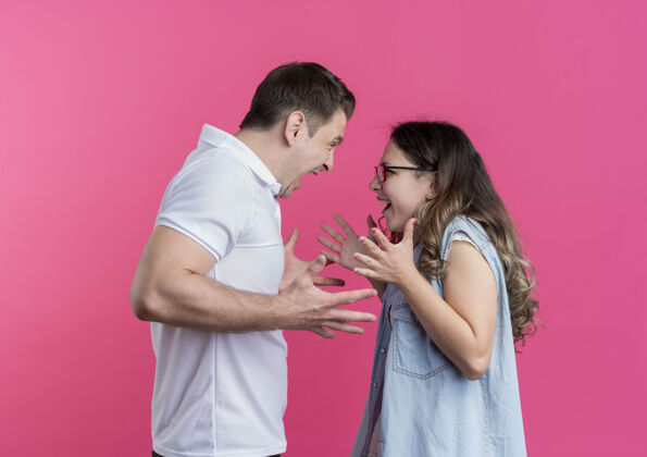年轻一对穿着休闲服的年轻夫妇站在粉红色的墙上吵架 互相喊叫各自衣服情侣