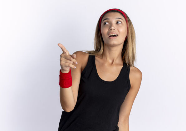 困惑身穿黑色运动服 头戴红色头巾的年轻健身女孩 用手指指着站在白墙上吓坏了的东西站健康年轻