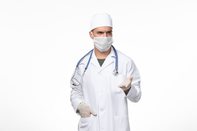 听诊器正面图：男医生穿着医疗服 戴着口罩抵御白色墙壁上的冠状病毒病大流行专业对抗查看