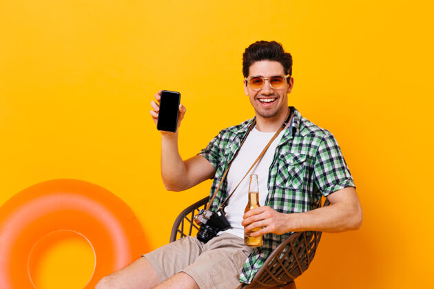 正面黑发男人微笑着展示他的智能手机穿着衬衫 t恤和短裤的男人坐在椅子上 手里拿着一瓶啤酒 放在橙色的空间里 还有一个充气的圆圈充气圈手机手机