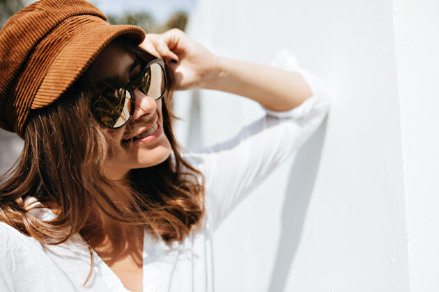 灌木戴着太阳镜的年轻女孩在街上摆姿势的特写镜头戴着棕色帽子微笑的女人靠在墙上放松休息城市
