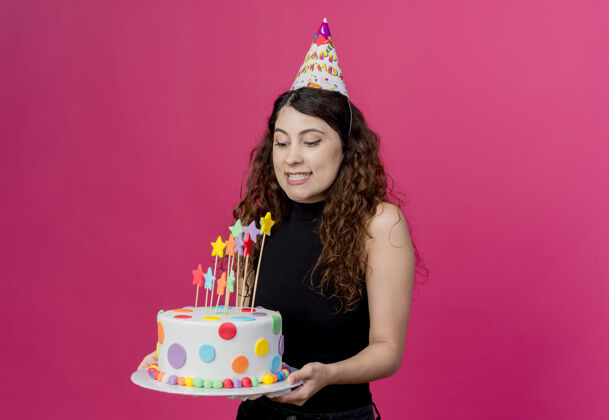 蛋糕年轻漂亮的女人 卷发 戴着节日帽 手里拿着生日蛋糕 站在粉色的墙上 快乐而积极的生日派对理念女人抱着年轻