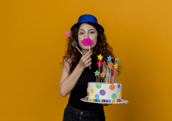 年轻年轻漂亮的女人 卷发 戴着派对帽 手里拿着生日蛋糕 手里拿着派对棒 快乐地站在橙色的墙上坚持女人帽子