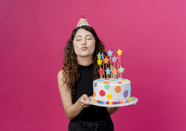抱着一个年轻漂亮的女人 卷发 戴着节日帽 手里拿着生日蛋糕 站在粉色的墙上 吹着生日派对的飞吻年轻吹蛋糕