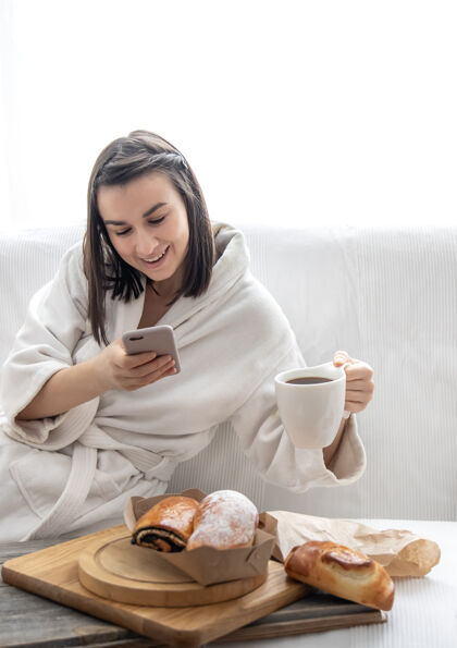 视频通话一个可爱的年轻女子穿着长袍坐在沙发上 为面包和咖啡拍照早午餐和周末概念周末早午餐早上