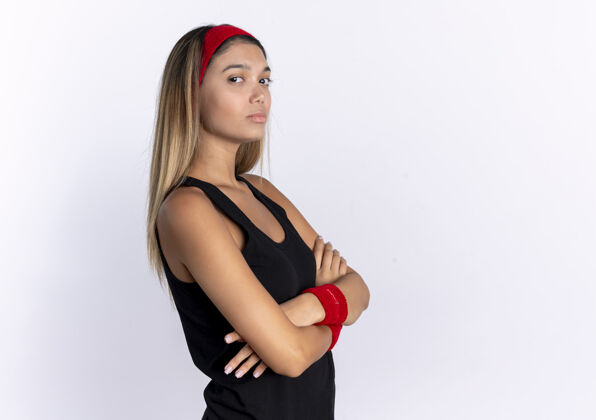 手臂身穿黑色运动服 头戴红色头带 双臂交叉 表情严肃自信的年轻健身女孩站在白色的墙上头带健康站立