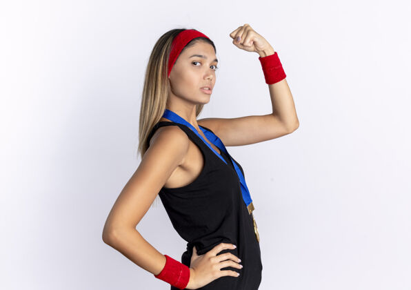 站身穿黑色运动服 头戴红色头带 脖子上戴着金牌的年轻健身女孩站在白色的墙上举起拳头 看上去很自信运动装信心周围