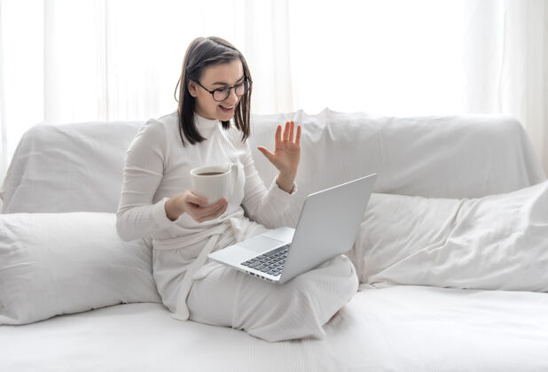 房子一个可爱的年轻女子正坐在家里的白色沙发上 穿着白色的裙子 坐在笔记本电脑前远程工作和自由职业者视频通话培训学生