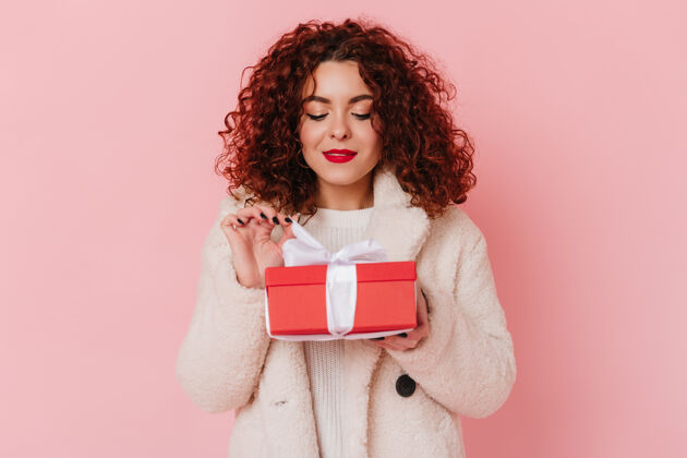红色唇膏迷人的女士拿着红色的礼盒 白色的缎带放在粉色的空间里卷发女孩的快照 穿着浅羊毛的衣服优雅羊毛表情