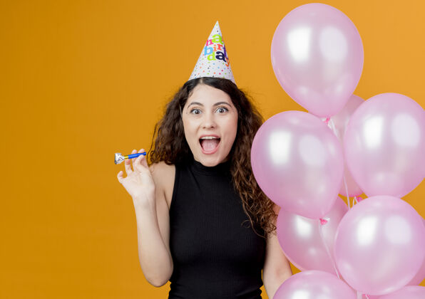 空气一个年轻漂亮的女人 卷发 戴着节日帽 手里拿着气球 吹着口哨 快乐而兴奋地微笑着 站在橙色的墙上 生日派对的概念站着女人兴奋