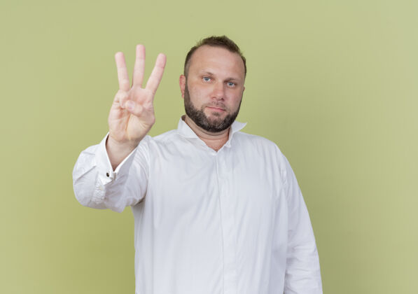 穿一个留着胡子的男人穿着白衬衫 手指着三号 表情严肃地站在轻质的墙上数字脸光