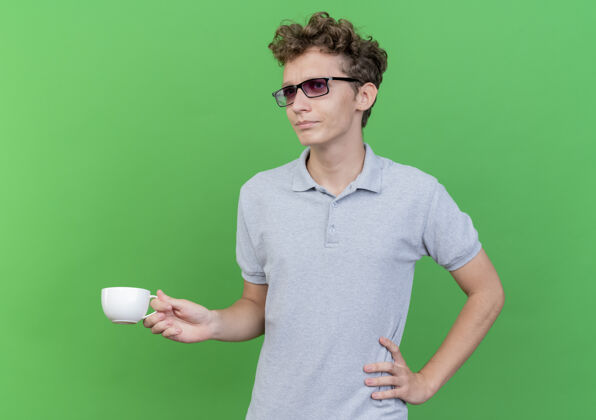 男人一个戴着黑眼镜 穿着灰色马球衫 拿着咖啡杯的年轻人站在绿色的墙上 一边看一边感到困惑和不快戴着不高兴马球