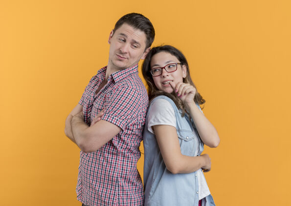 微笑一对穿着休闲服的年轻夫妇背靠背地站在橙色的墙上微笑着背部站立衣服