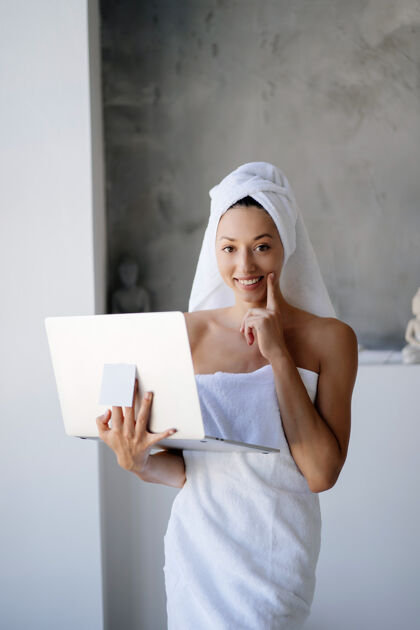 毛巾穿着白毛巾的女自由职业者站在浴室里拿着笔记本电脑自由职业者笔记本电脑白色