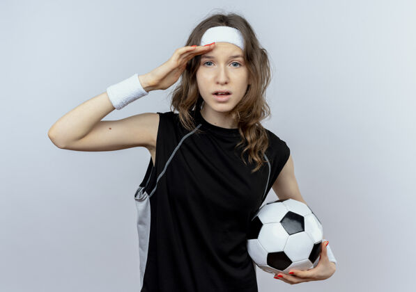 远穿着黑色运动服的年轻健身女孩 头上带着足球 手举着头站在白色的墙上遥望远方看站头
