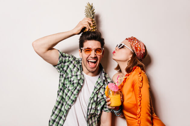 放松一个戴着明亮太阳镜的快乐的黑发男人把菠萝放在头上 他的女朋友亲吻他的脸颊大笑漂亮情侣