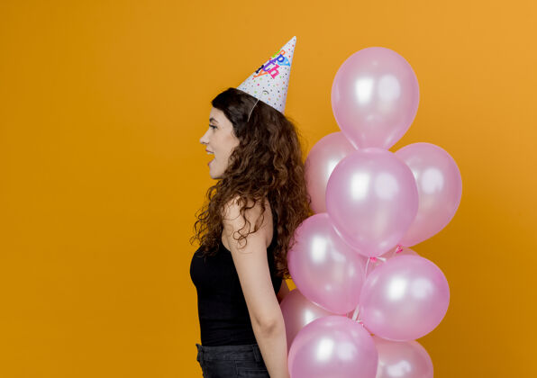 年轻一个年轻漂亮的女人 卷发 戴着节日帽 手持气球 站在橙色的墙上庆祝生日气球空气侧着